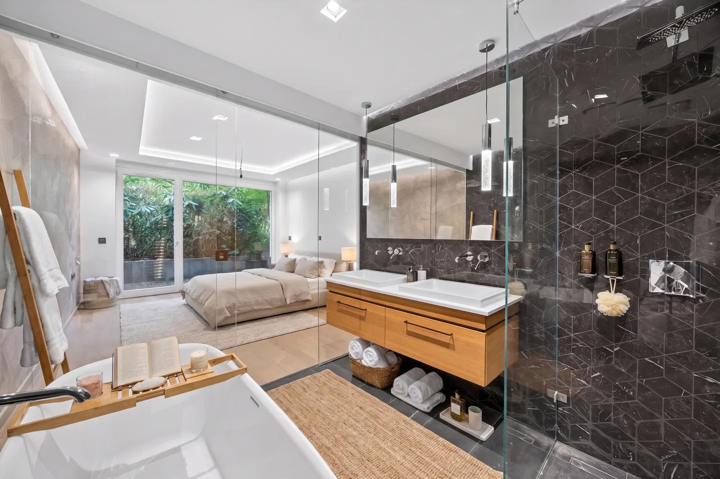A modern bathroom with a glass wall to the bedroom, featuring a tub, double vanity, and marble shower.