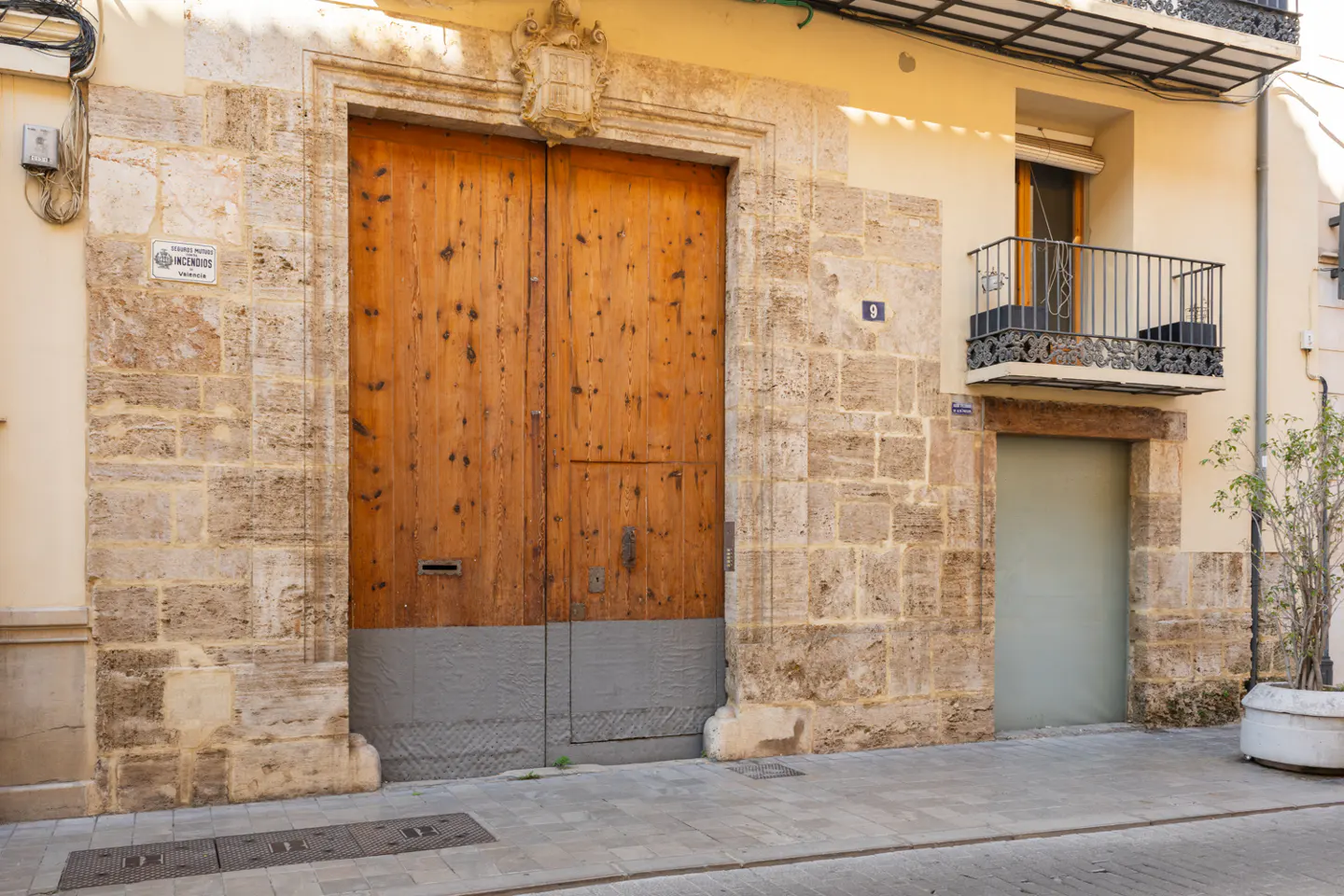 Street view of a building with a large wooden door and stone facade. A small balcony is visible above a smaller door.