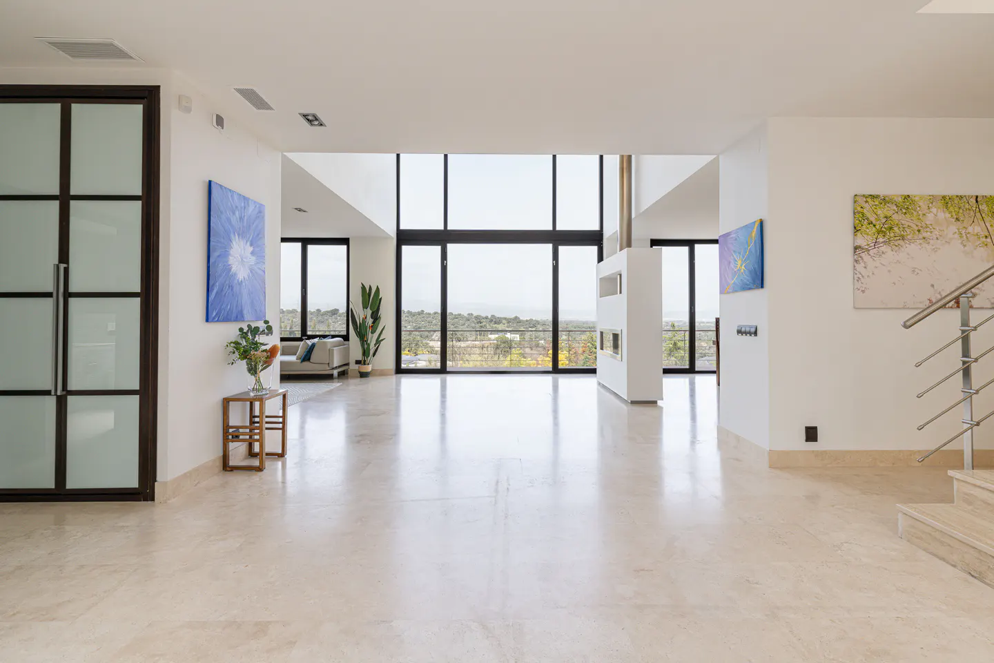 Bright, modern home interior with marble floors, white walls, and large windows. Artwork adorns the walls, and a staircase is visible.
