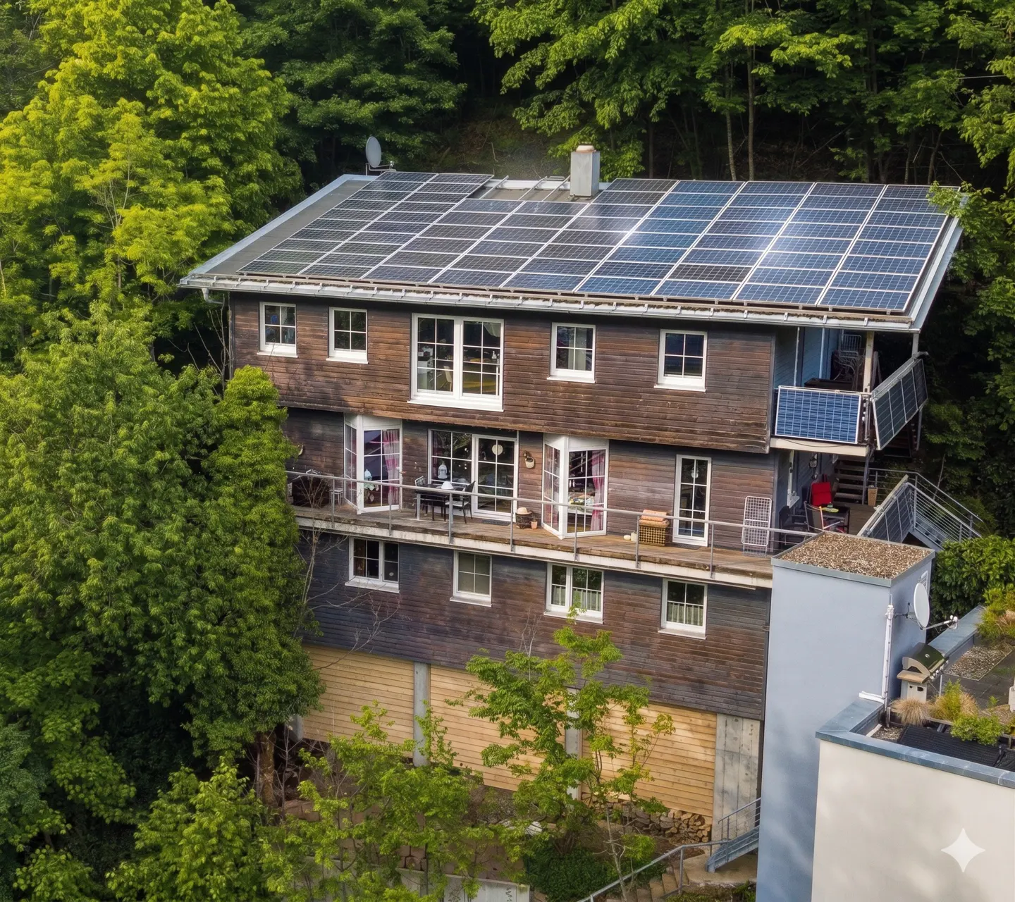 Three-story brown house with solar panels on the roof, surrounded by green trees. Balconies with furniture are visible.