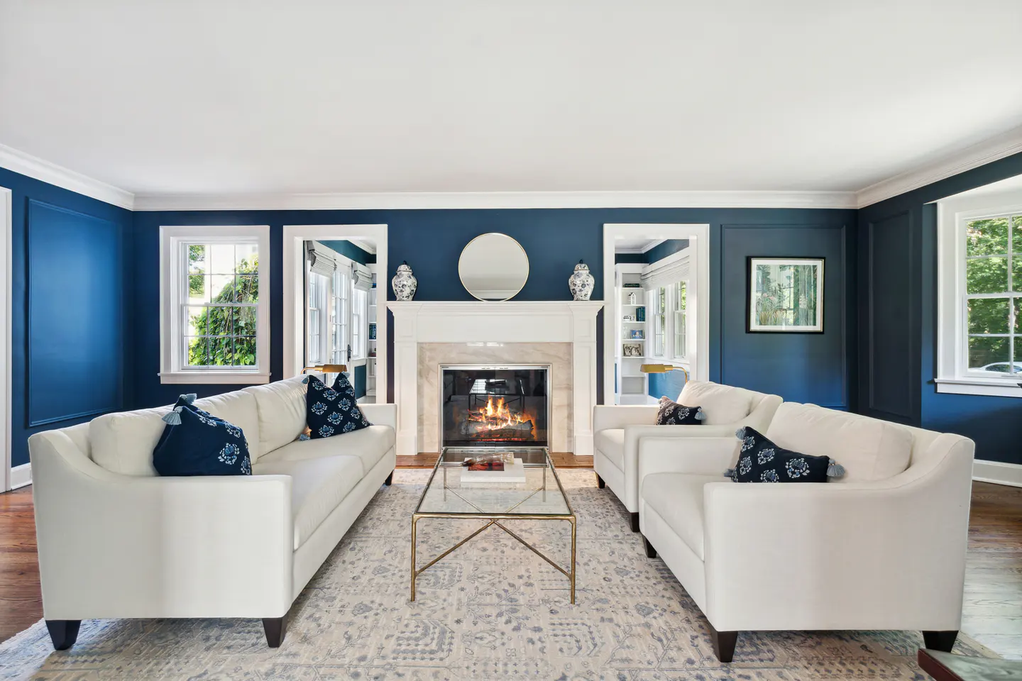 Living room with blue walls, white sofas, and a fireplace with a round mirror above it. A glass coffee table sits on a patterned rug.