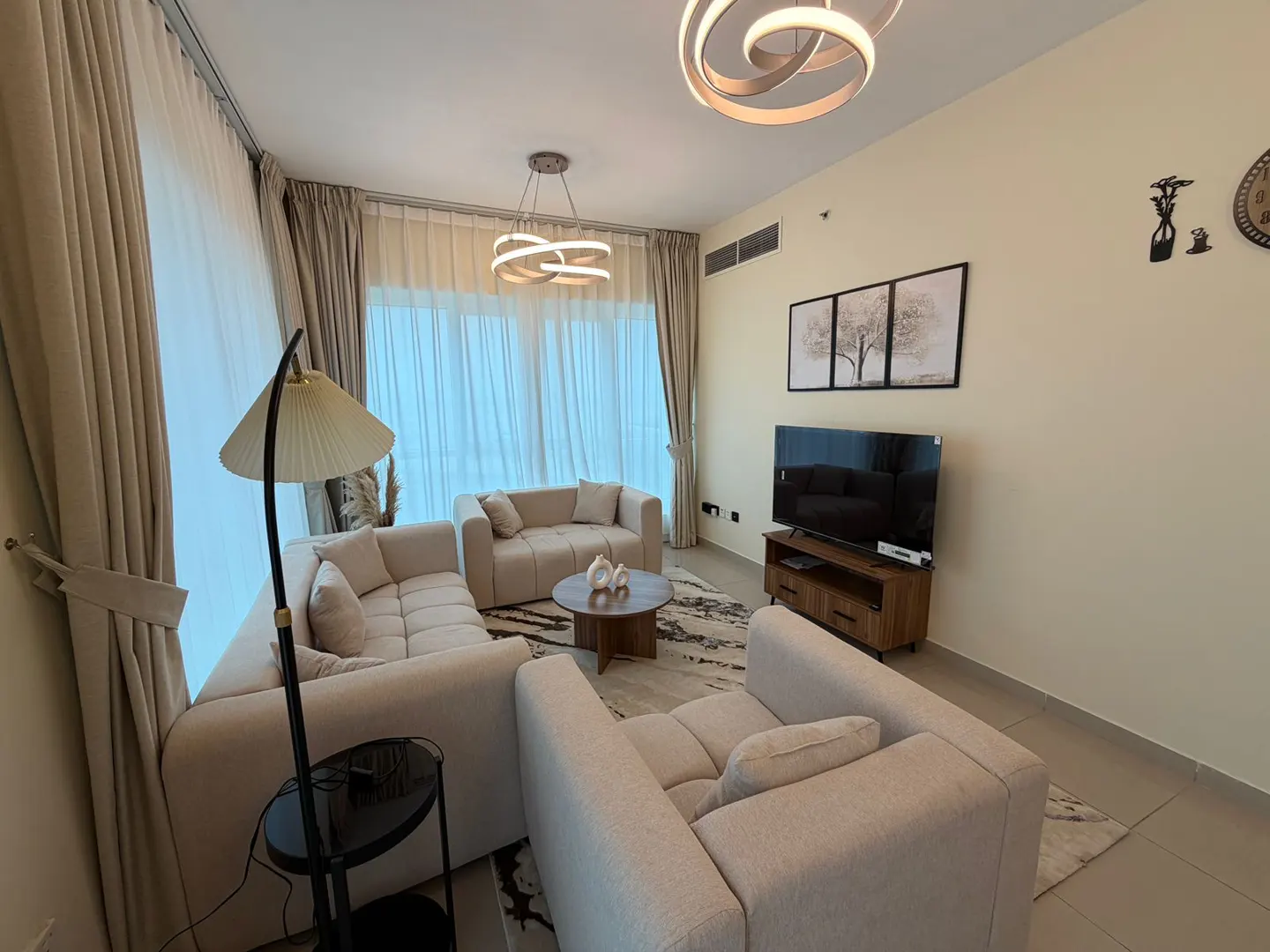 Living room with beige sofas, round wood table, TV, and modern lighting fixtures. Neutral color palette.