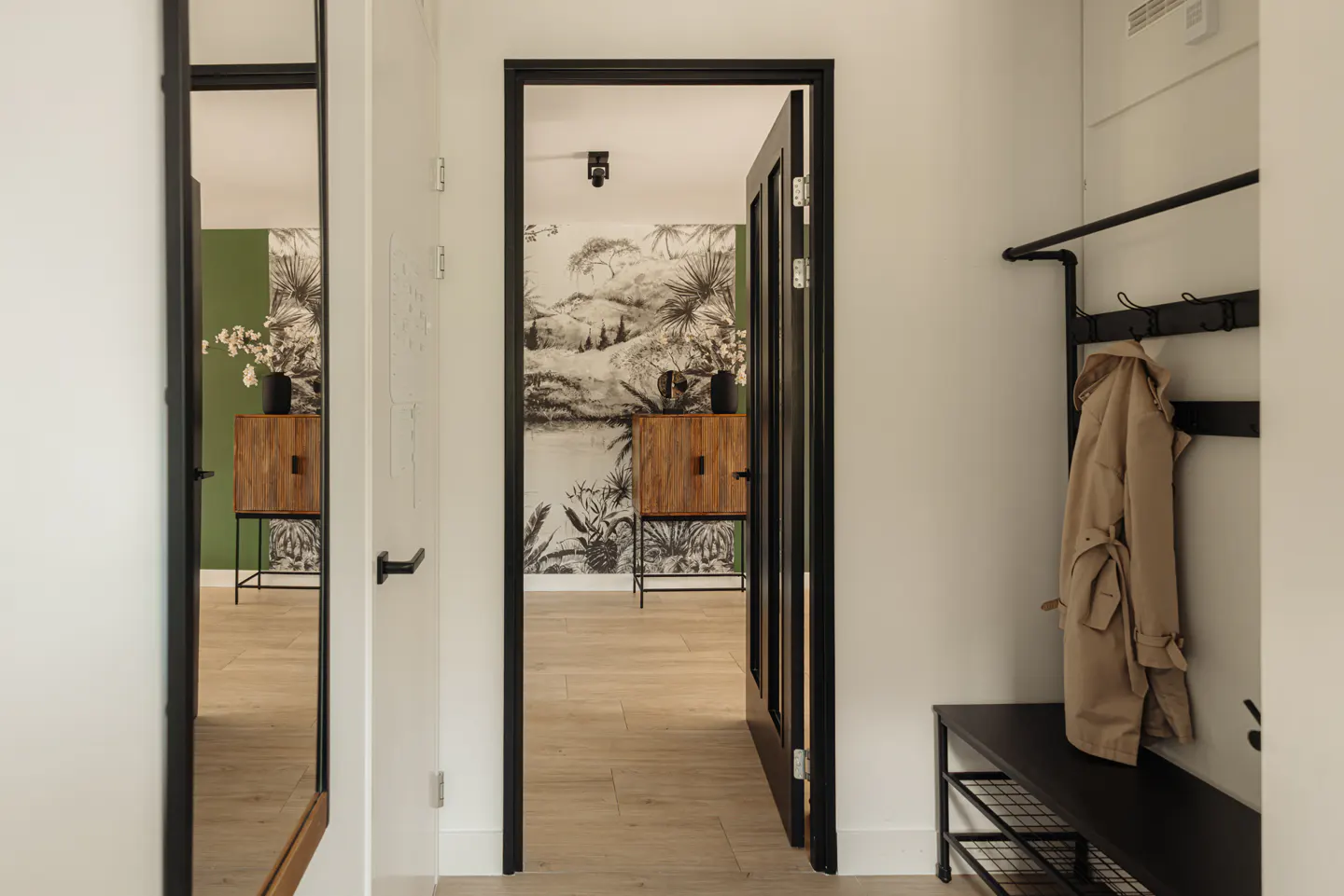 A bright hallway with white walls, a black-framed mirror, and an open door revealing a room with a wooden cabinet and jungle-themed wallpaper. A coat hangs on a black rack.