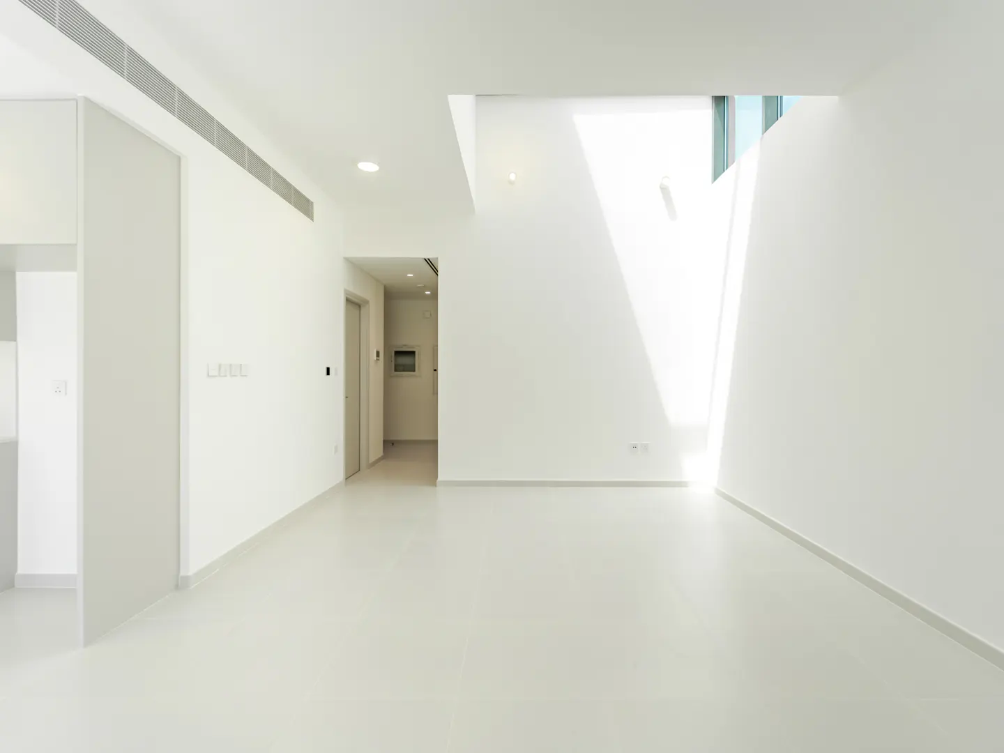 Bright, empty room with white walls and floors. Sunlight streams in from a high window. A doorway leads to another room.