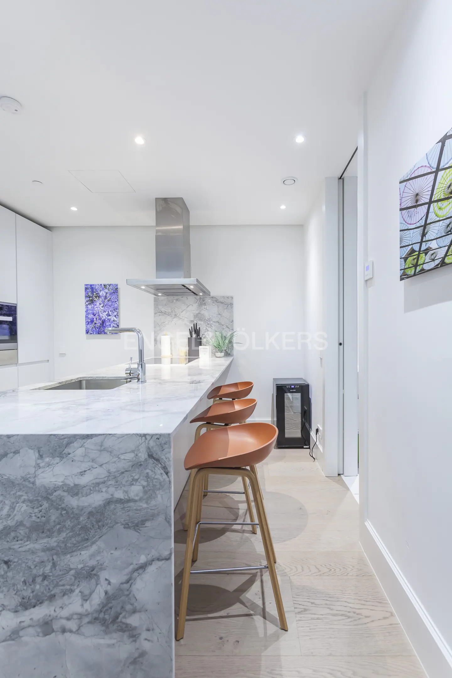 Modern kitchen with marble island, stainless steel hood, and three brown bar stools. White walls and light wood floors.