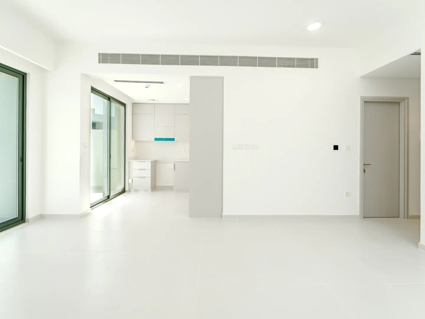 Bright, empty room with white walls, floor, and kitchen. Sliding glass doors lead outside. A gray door is on the right.
