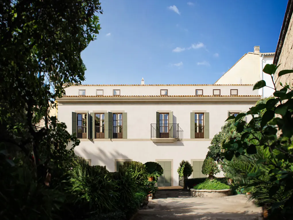 Two-story white building with green shutters, framed by lush greenery under a blue sky. Balconies accent the upper level.