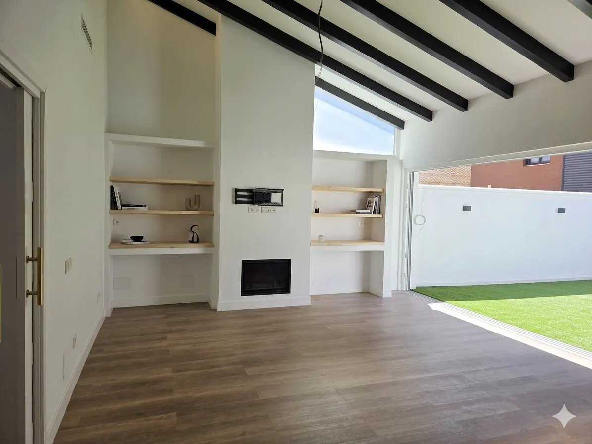 Bright living room with wood floors, white walls, and black ceiling beams. Built-in shelves flank a fireplace. Sliding glass doors open to a green lawn.