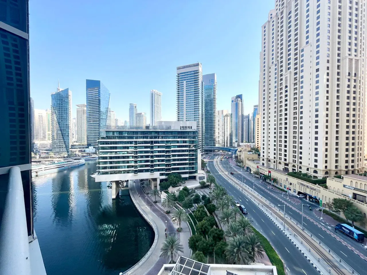 View of Dubai Marina with skyscrapers, a canal, and a highway. Palm trees line the walkway. Blue sky.