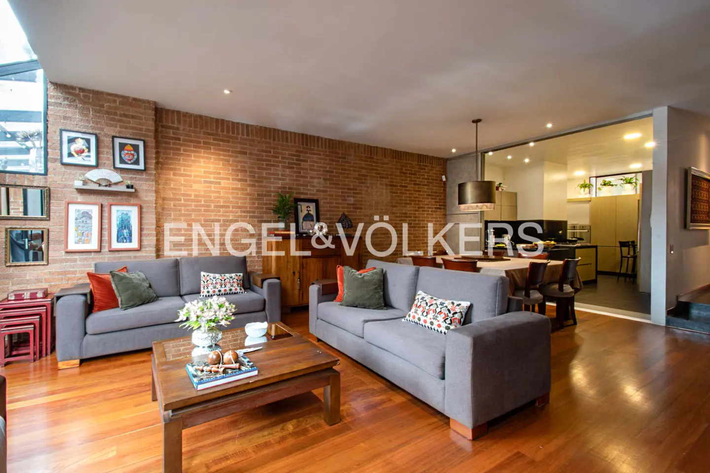 Living room with gray sofas, wood floors, and brick wall. A wood coffee table sits in front of the sofas. Engel & Völkers logo is visible.