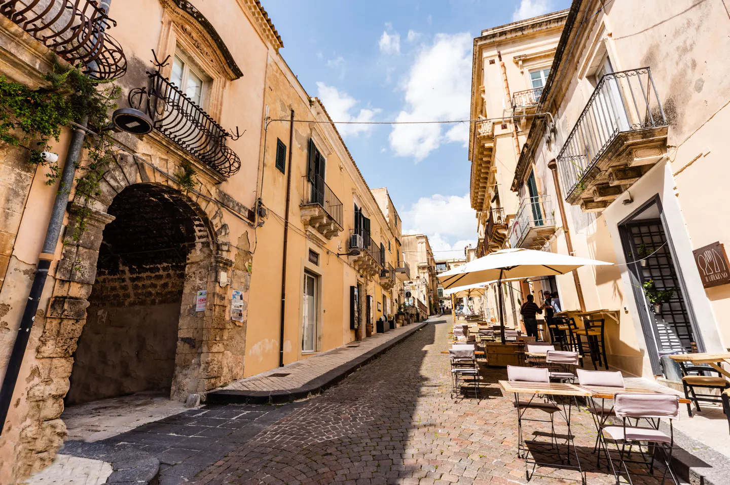 Cobblestone street in Italy with peach buildings, iron balconies, and outdoor cafe seating under white umbrellas.