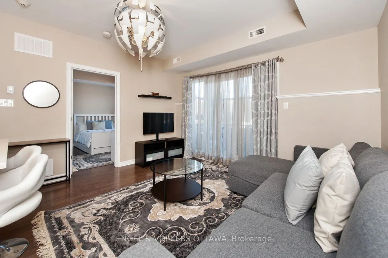 A living room with a gray sofa, patterned rug, and a view into a bedroom with a white bed. A modern light fixture hangs from the ceiling.