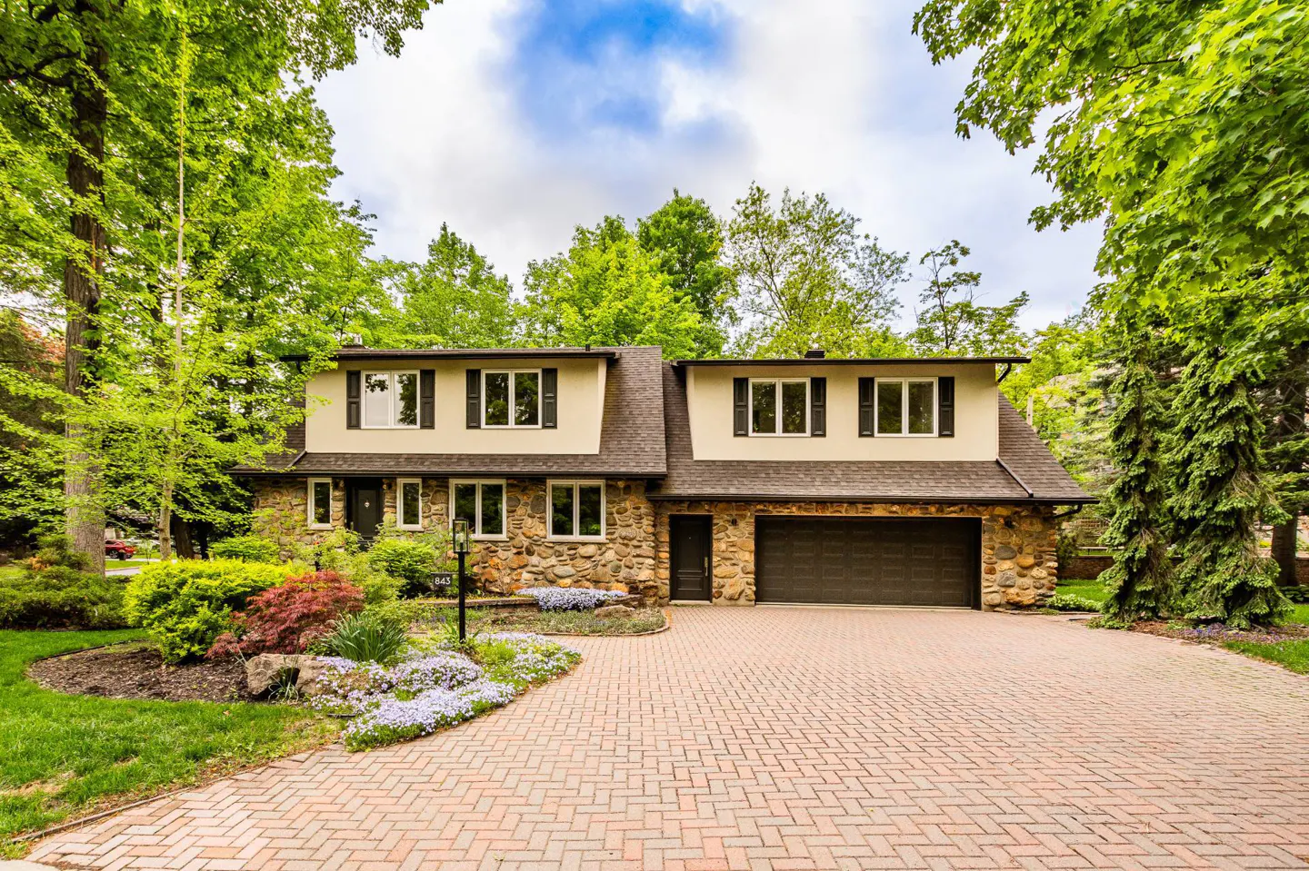 Two-story home with stone lower level, tan upper level, and a brick driveway surrounded by lush green trees and flowers.