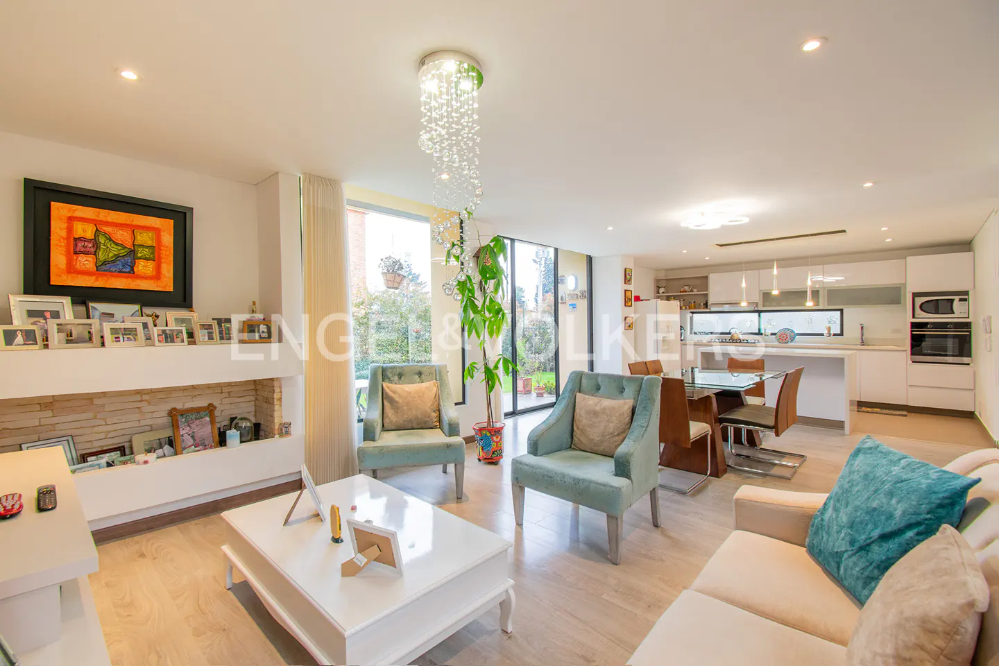 Bright, open-plan living room with light wood floors, white walls, and a modern chandelier. Turquoise armchairs and a beige sofa add color. Kitchen in the background.