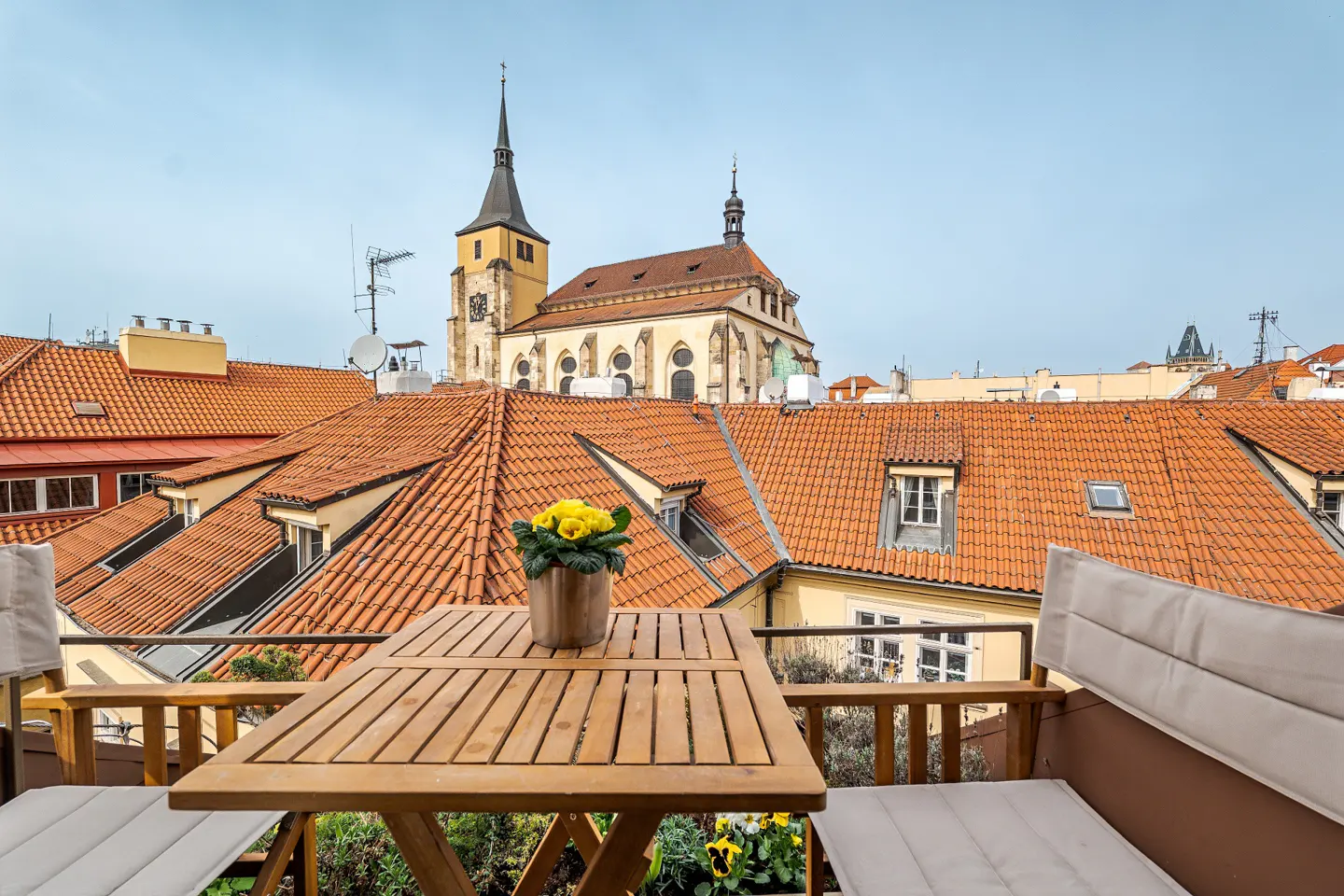 Balcony view of Prague rooftops. A wooden table with yellow flowers sits in front of orange tile roofs and a church steeple.