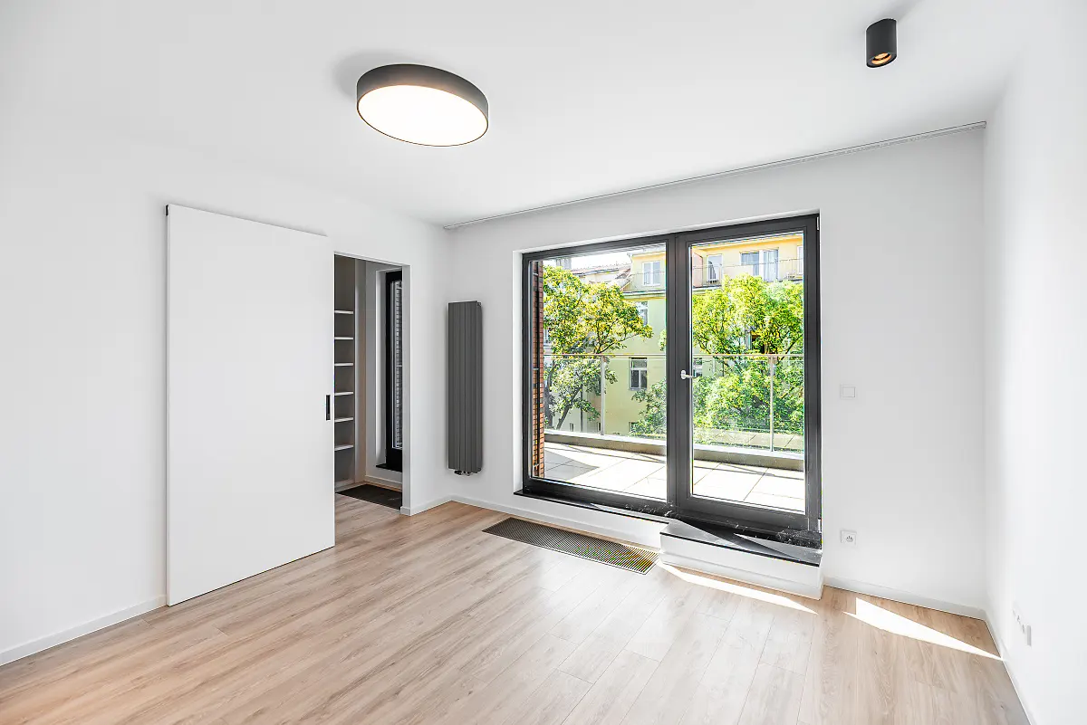 Bright, minimalist room with light wood floors, a sliding door to a closet, and a balcony door overlooking green trees.