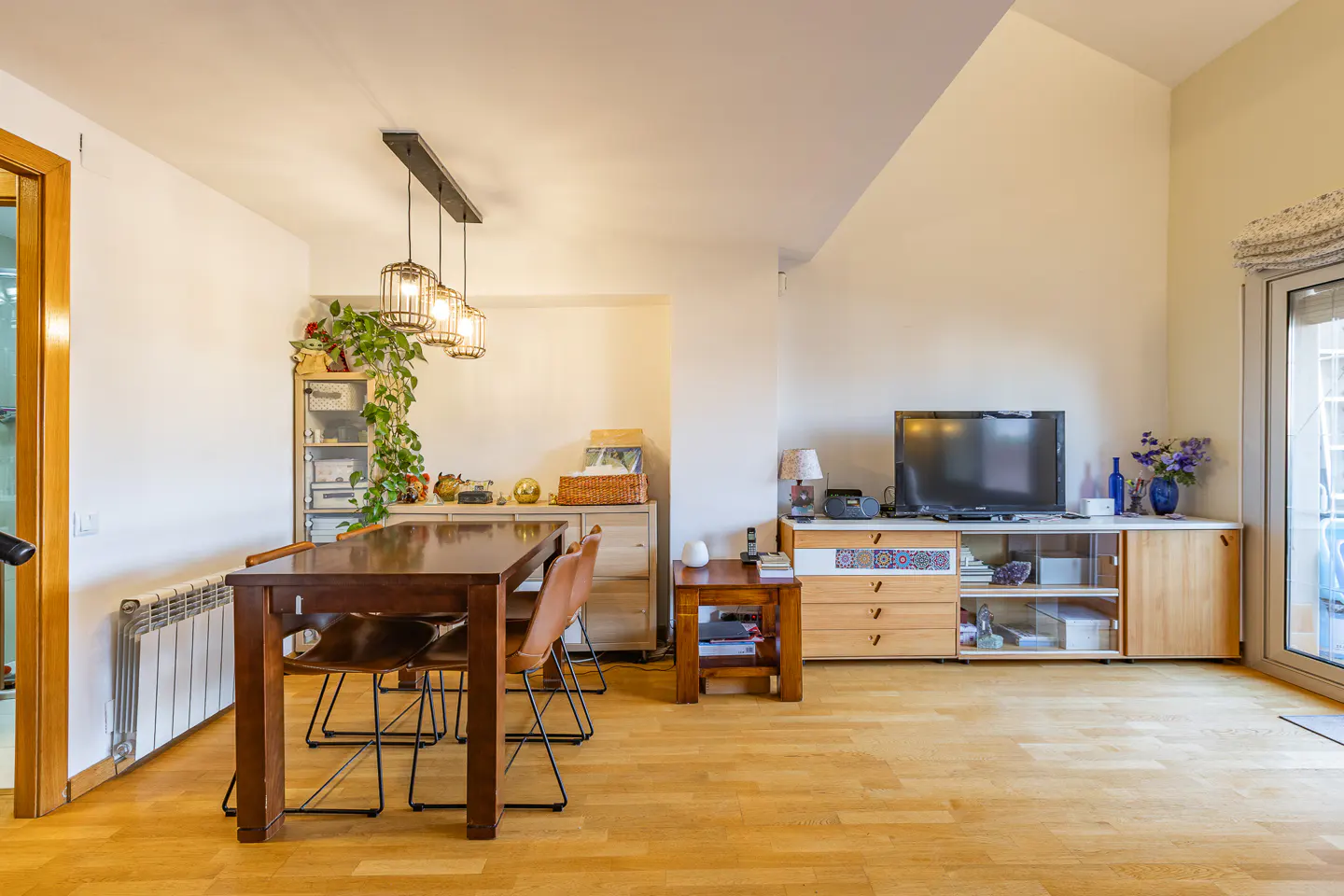 Bright, modern living room with wood floors, a dining table with chairs, and a TV on a cabinet. Three pendant lights hang above the table.