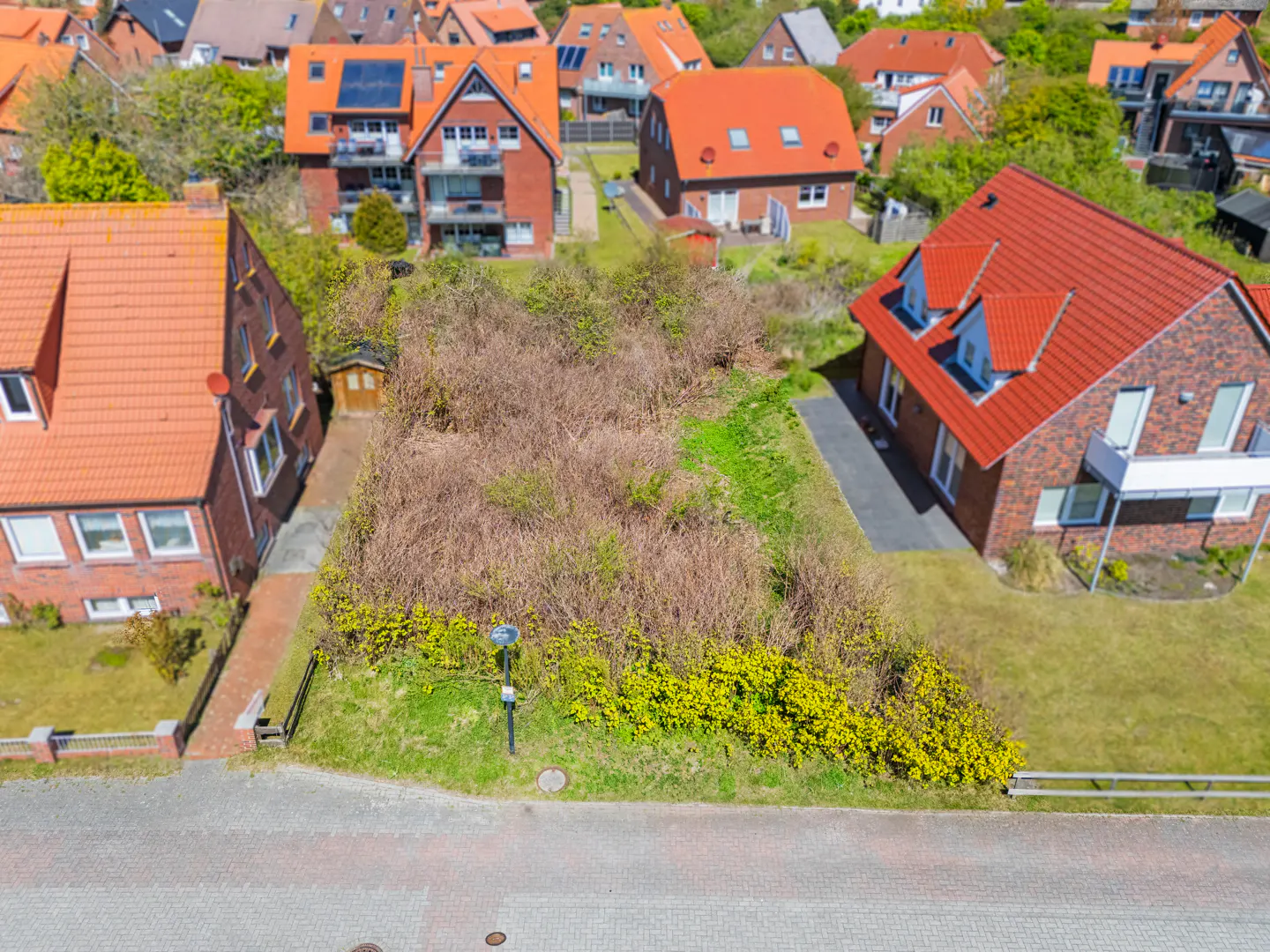 Aerial view of a vacant lot overgrown with brush, surrounded by red-roofed houses in a residential neighborhood.