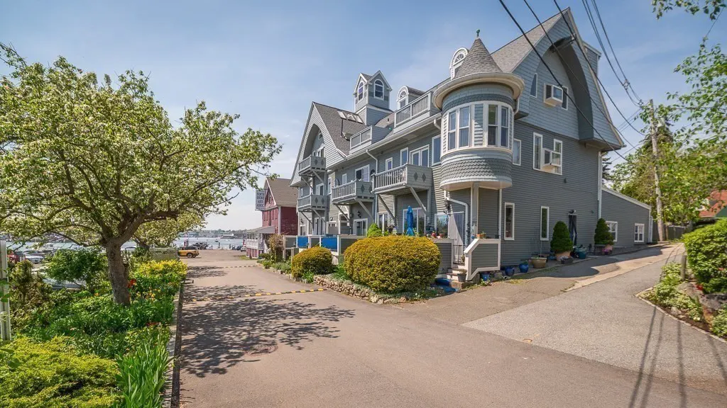 A multi-story grey Victorian-style building with a turret, balconies, and lush landscaping along a paved driveway.