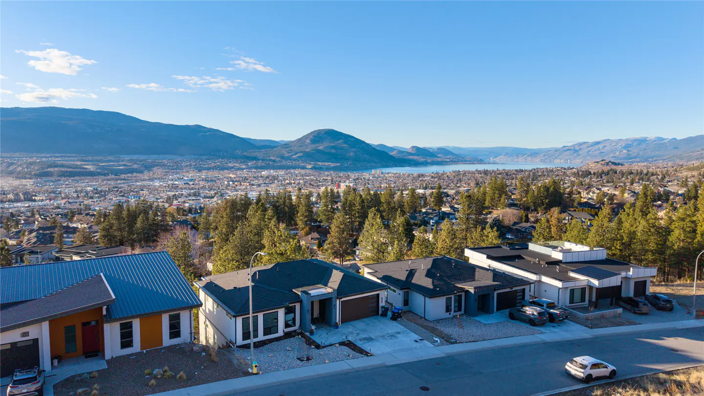 Aerial view of modern homes on a hill overlooking a city, lake, and mountains under a clear blue sky.