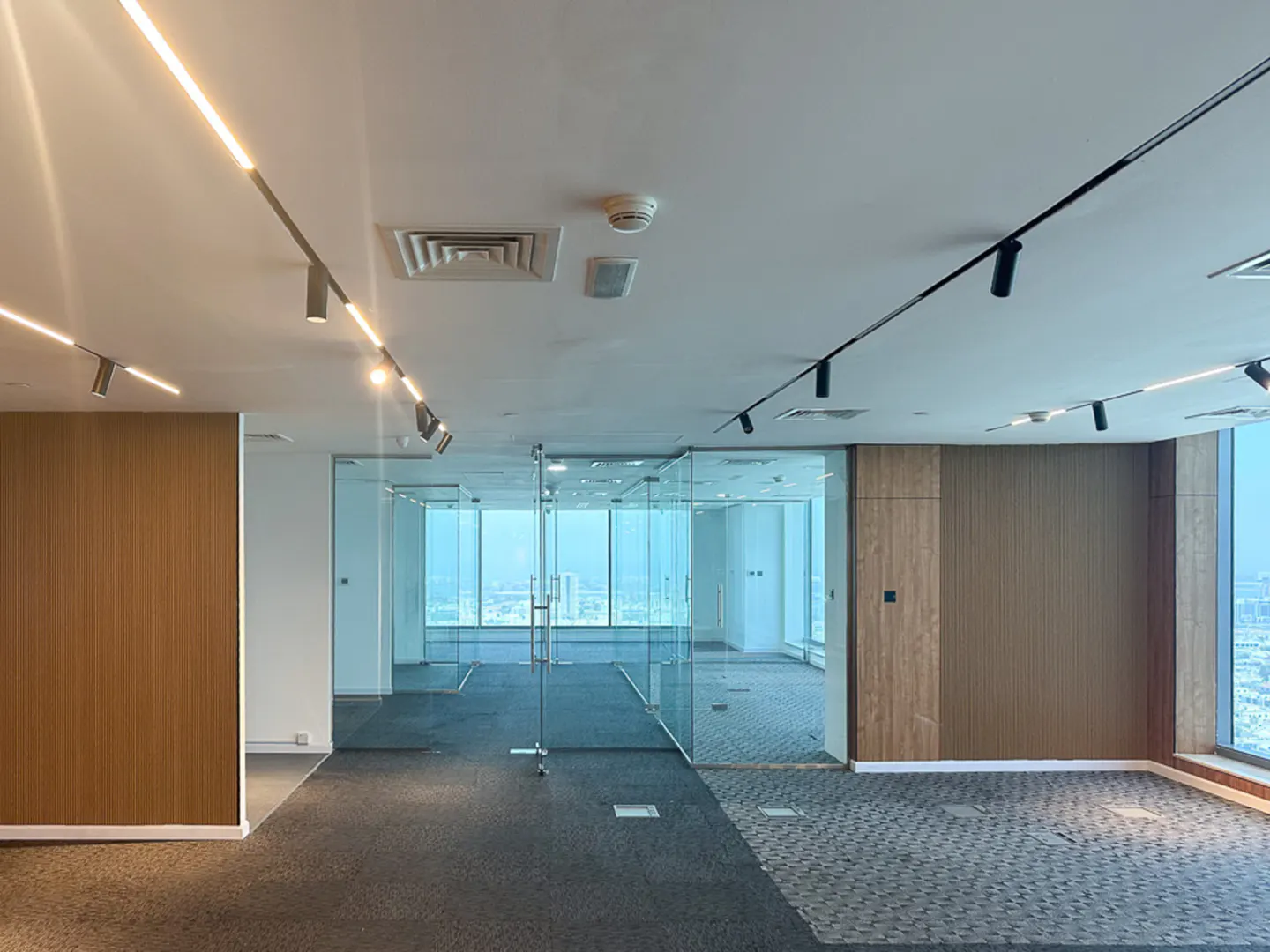 An empty office space with glass walls, gray carpet, and wood paneling. Track lighting illuminates the room with a city view visible through the windows.