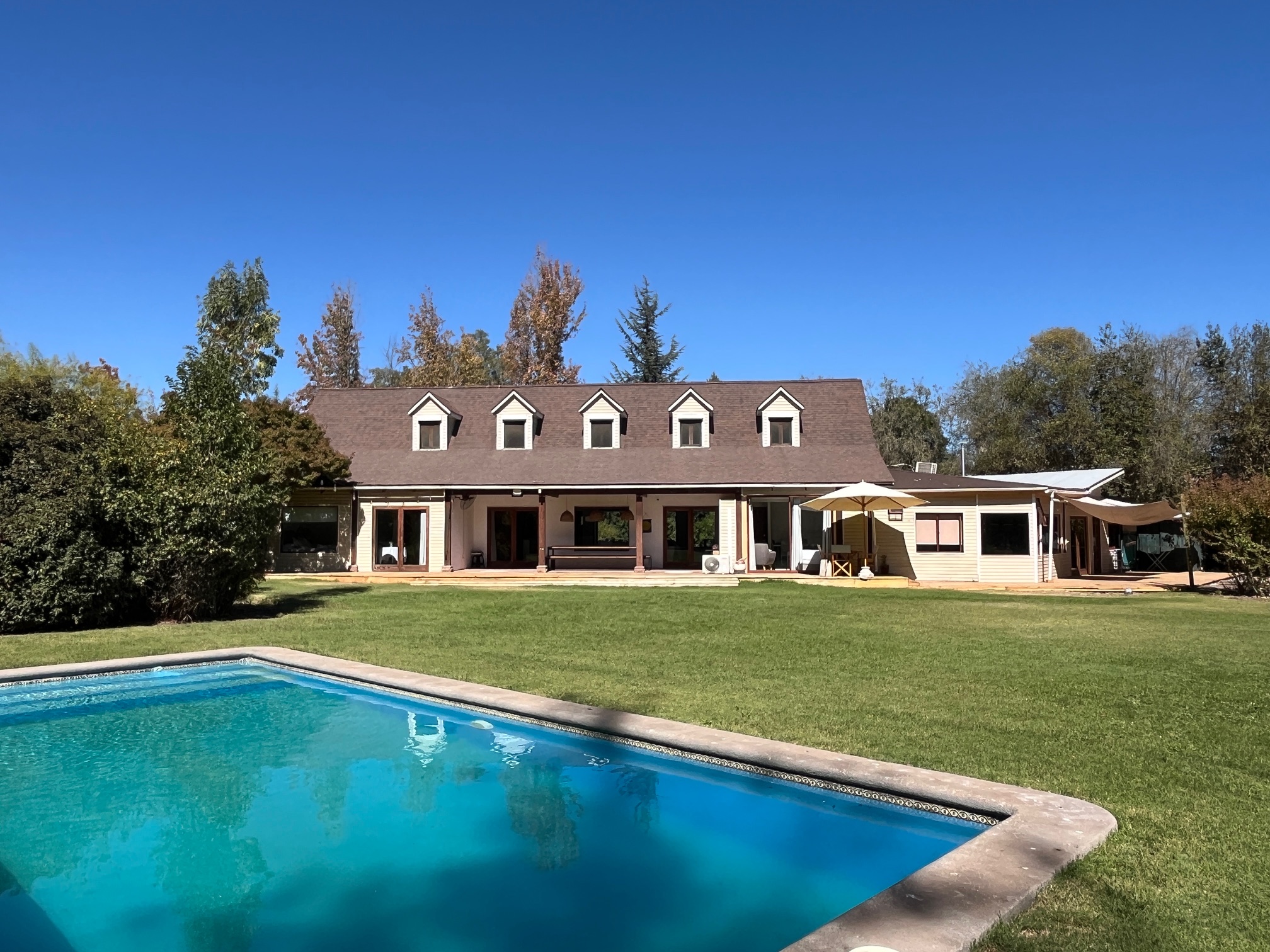 A large house with a brown roof and white walls overlooks a blue swimming pool and green lawn under a clear blue sky.