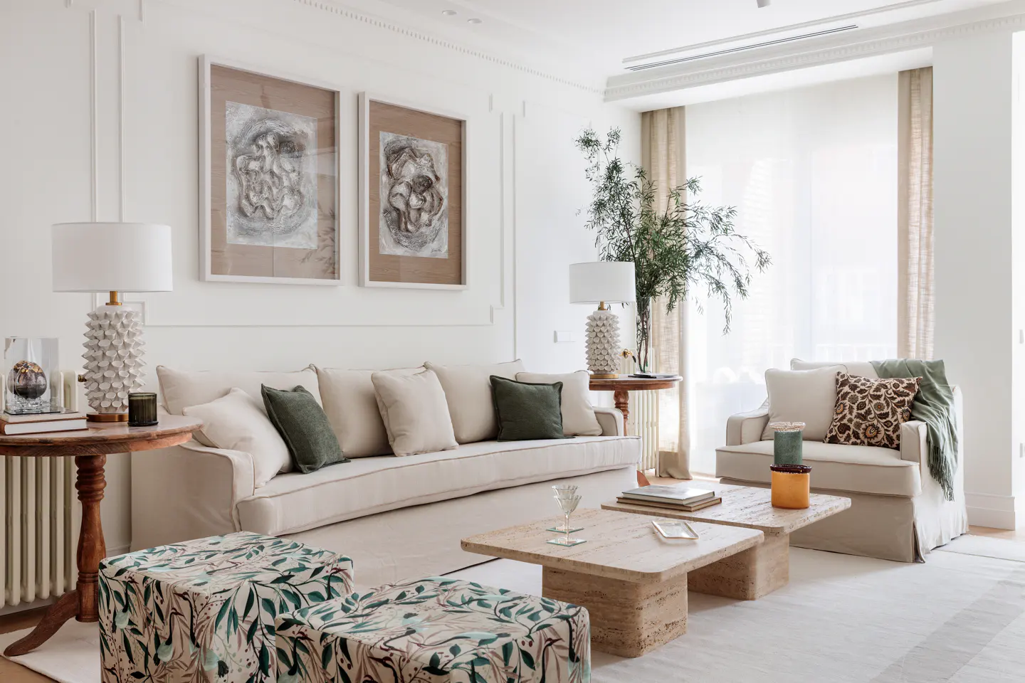 Bright living room with white walls, beige sofa, patterned ottomans, and travertine coffee tables. Artwork hangs above the sofa.