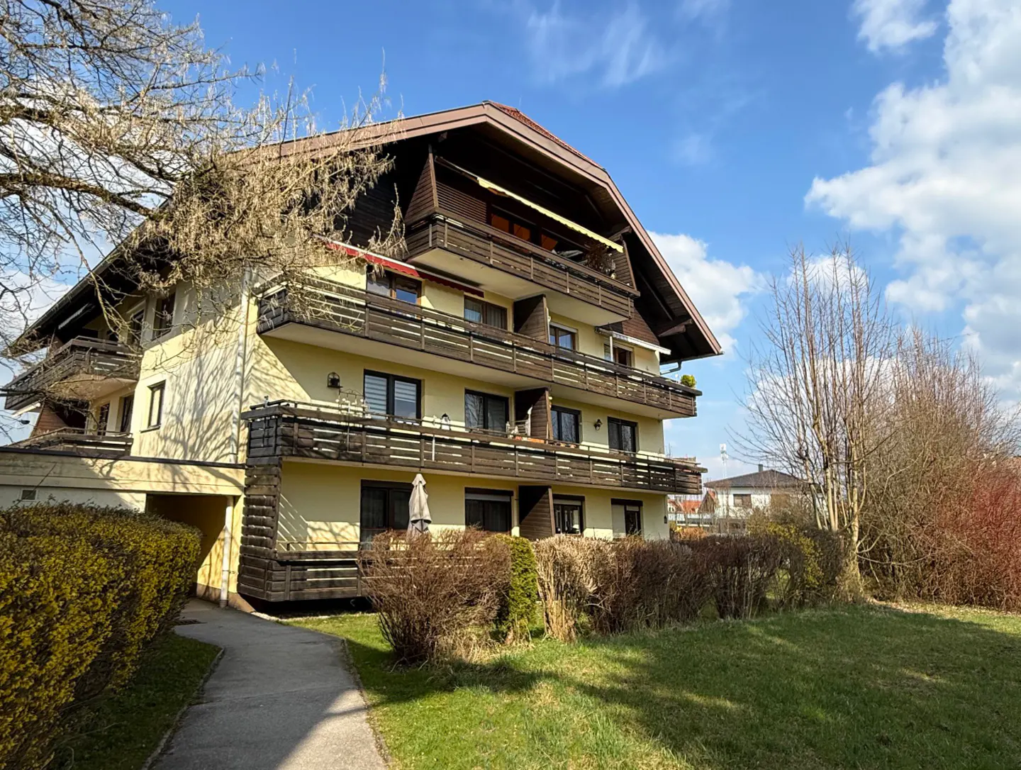 Three-story yellow apartment building with brown balconies and a brown roof, set against a blue sky with clouds.