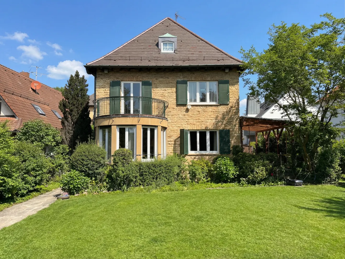 Two-story tan brick house with green shutters, a brown roof, and a green lawn on a sunny day.