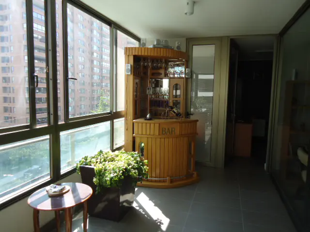 Sunroom with a wooden bar, plants, and a small table. Large windows offer a city view.