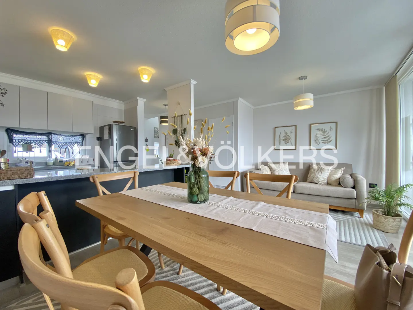 Open-concept living space with a wooden dining table, chairs, and a floral centerpiece. A gray sofa and fern art are in the background.