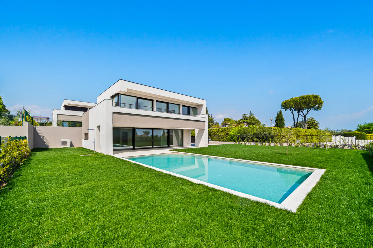 Modern two-story house with a rectangular pool and green lawn under a clear blue sky.