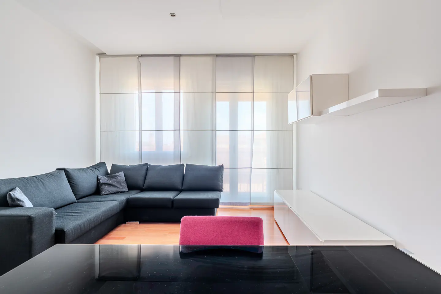 Bright living room with a black sectional sofa, pink ottoman, and white shelving against a large window.