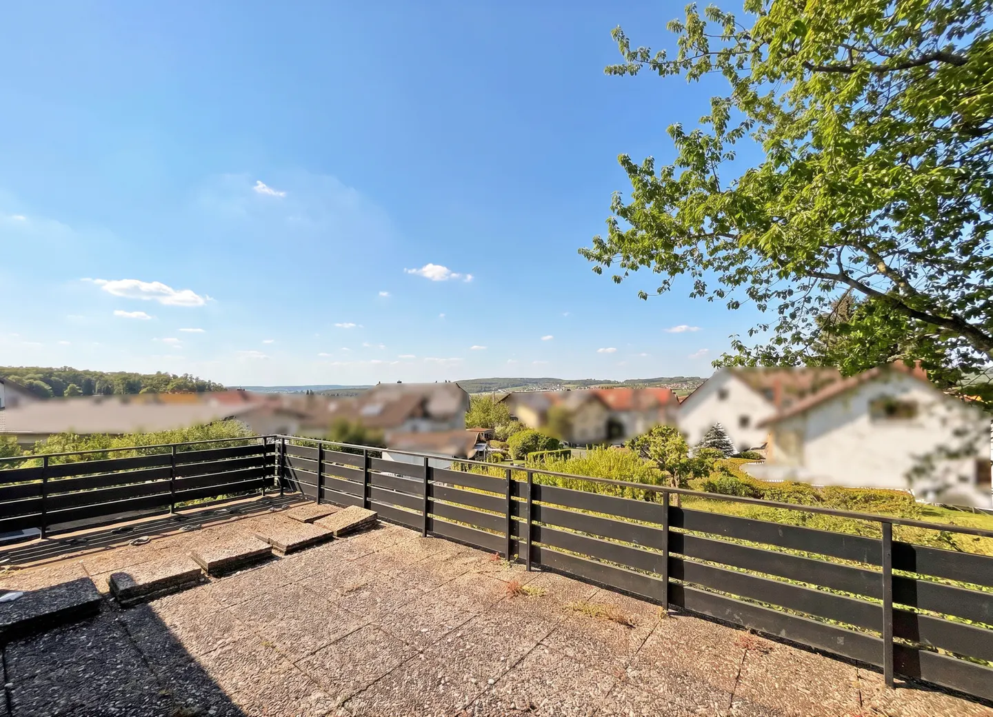 View from a concrete patio with a black fence overlooking a town with trees under a blue sky.