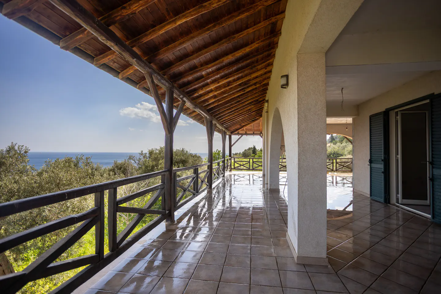 Covered patio with wood beams, tile floor, and dark wood railing overlooking the ocean and trees. Arched doorways lead inside.