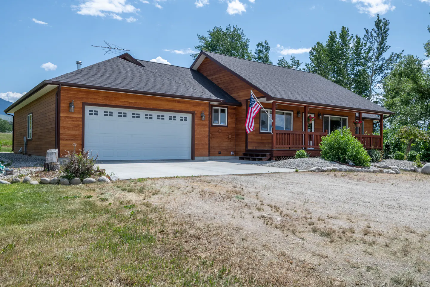 A single-story wood house with a gray roof, white garage door, and American flag on the porch.