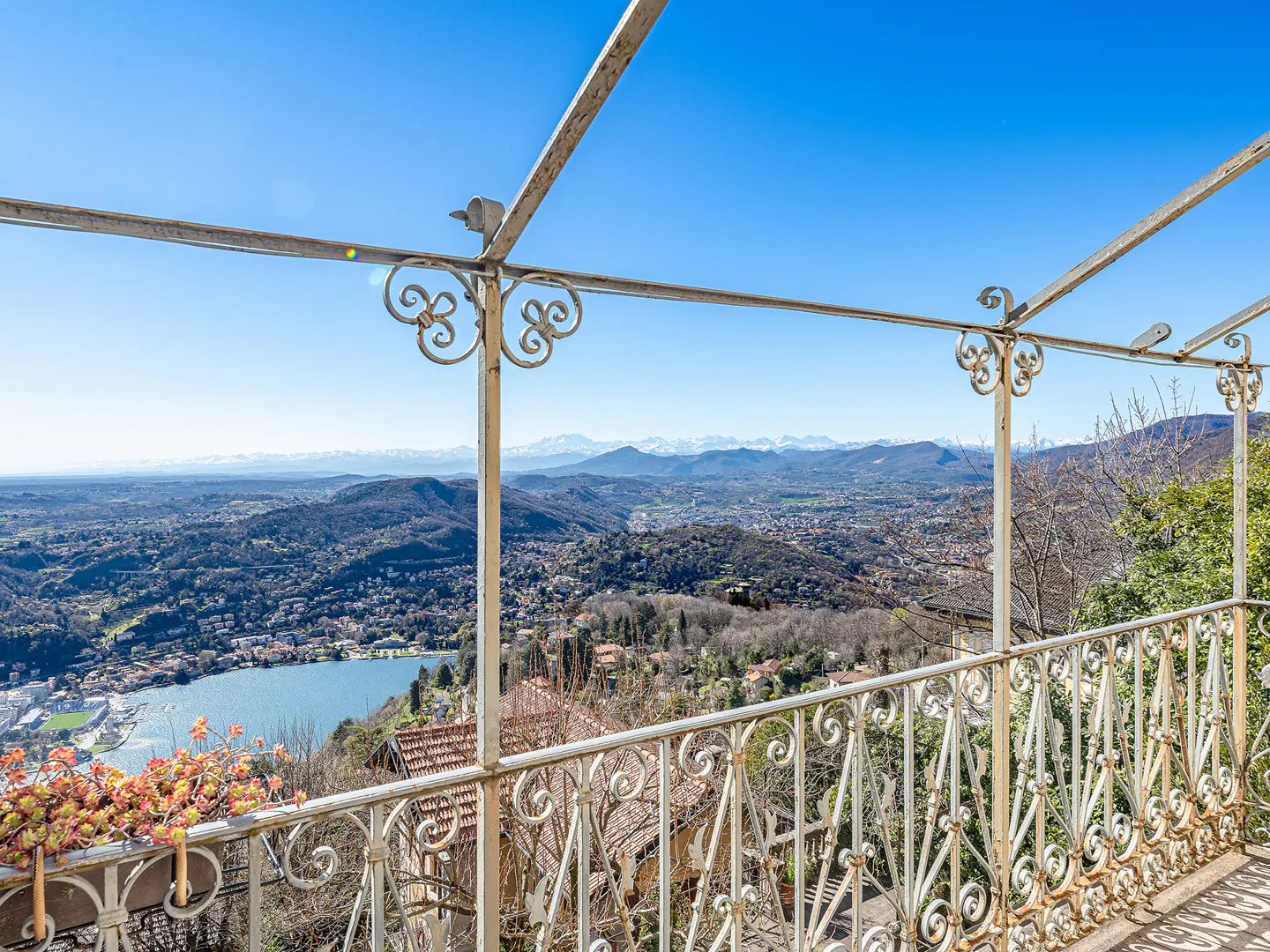 Scenic view from a balcony with white metal railings, overlooking a lake, hills, and distant snow-capped mountains under a clear blue sky.