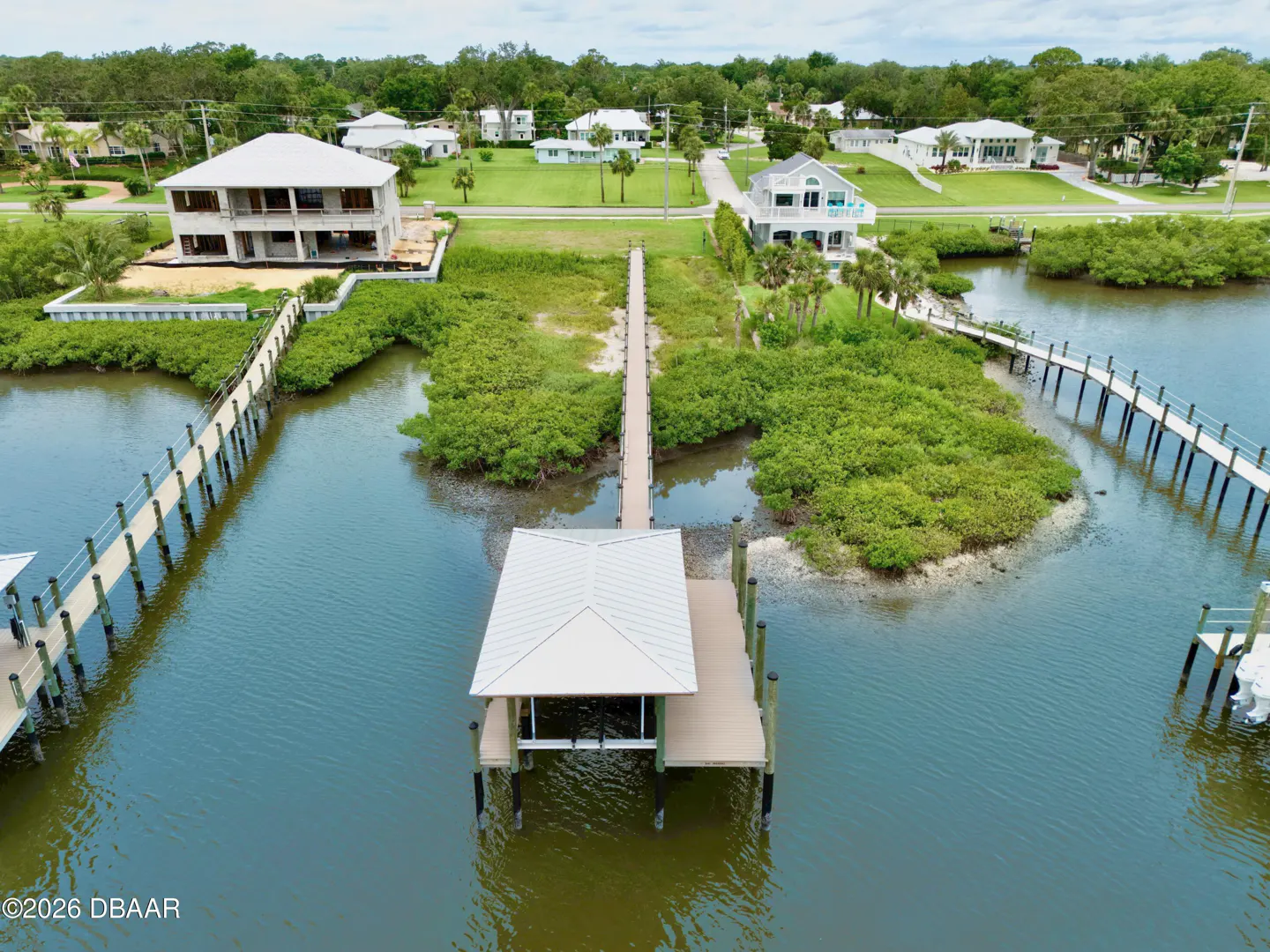 Aerial view of waterfront homes with docks on a sunny day. Green trees and blue water surround the properties.