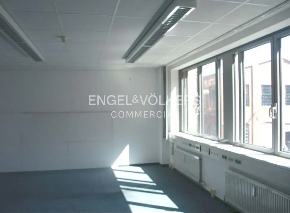 Bright, empty office space with large windows, white walls, and gray carpet. Engel & Völkers Commercial logo on the wall.