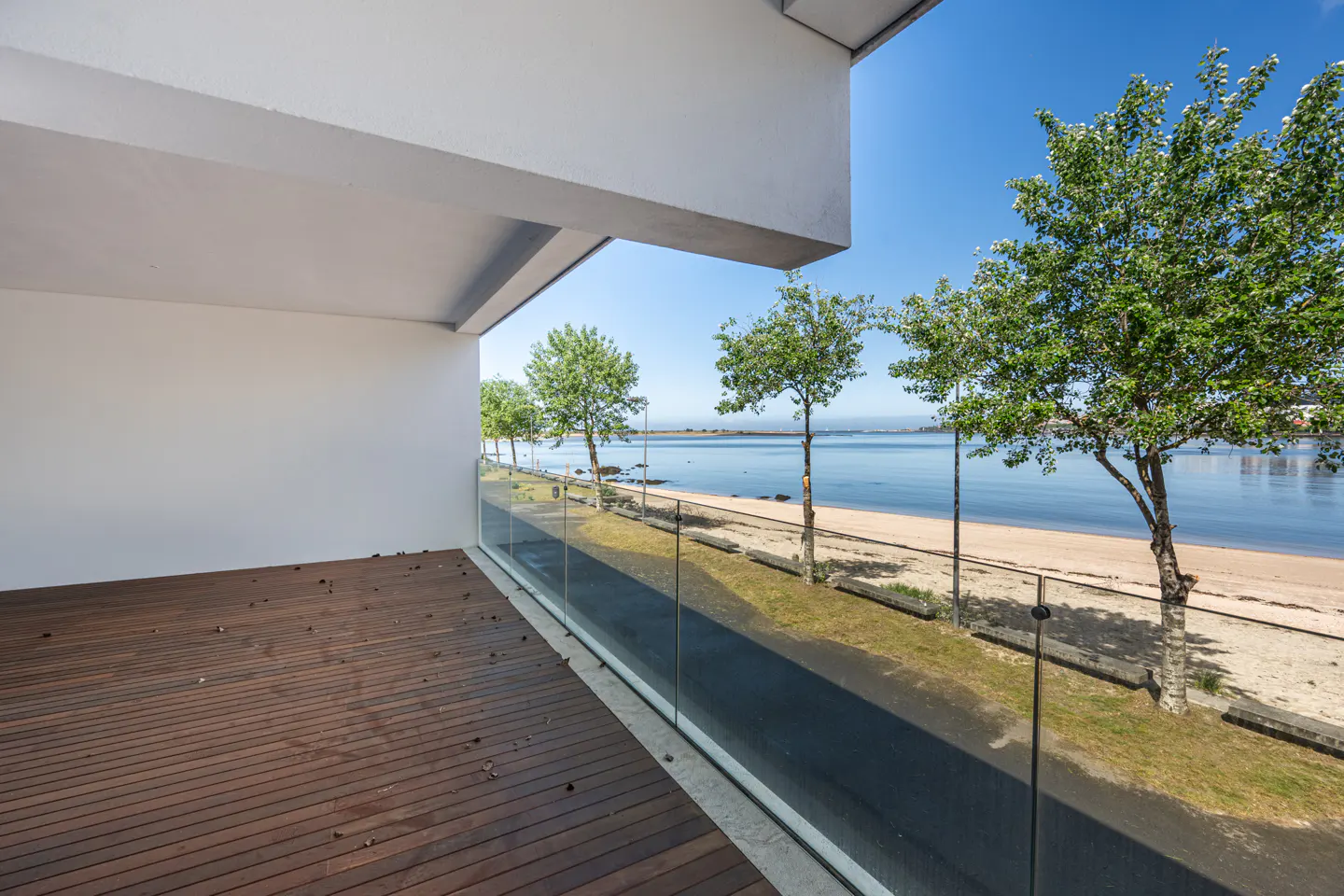 Balcony view of a beach. Brown wood floor, white walls, and a glass railing. Trees line the beach with blue water and sky.