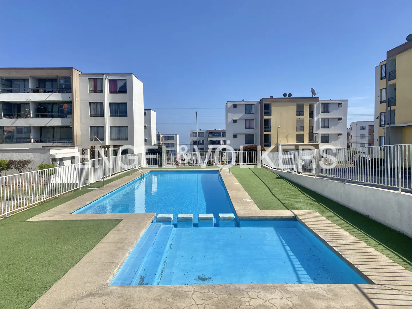 Outdoor pool with blue water, surrounded by artificial grass and white fence, with apartment buildings in the background.