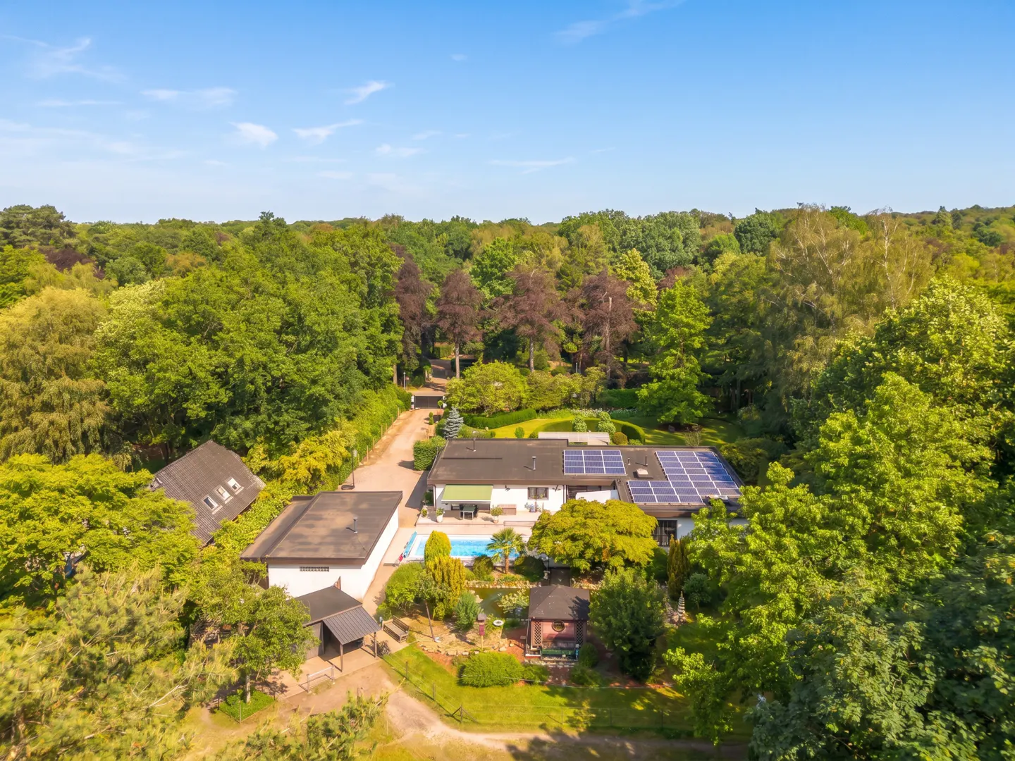 Aerial view of a white house with solar panels, a swimming pool, and a small gazebo, surrounded by lush green forest.