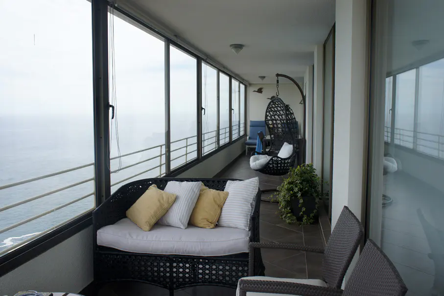 A sunroom with wicker furniture, including a sofa with pillows, chairs, and a hanging swing chair, overlooks the ocean through large windows.
