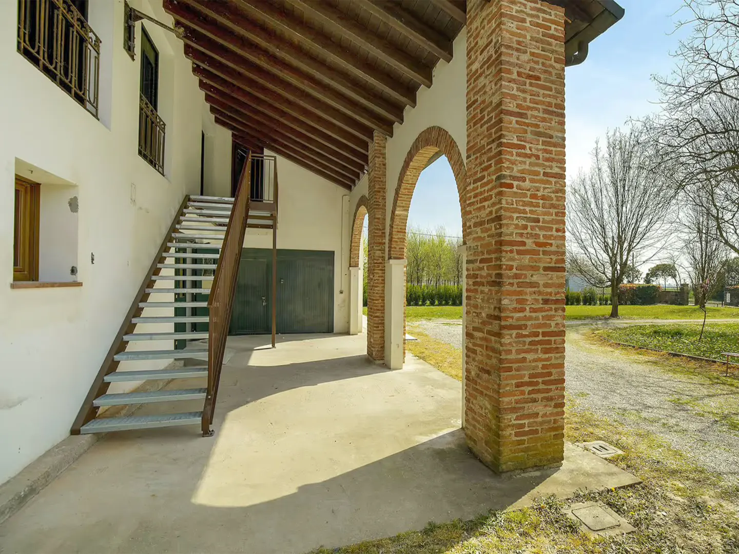 Exterior view of a white building with brick arches, a metal staircase, and a green garage door. The ceiling has exposed wooden beams.