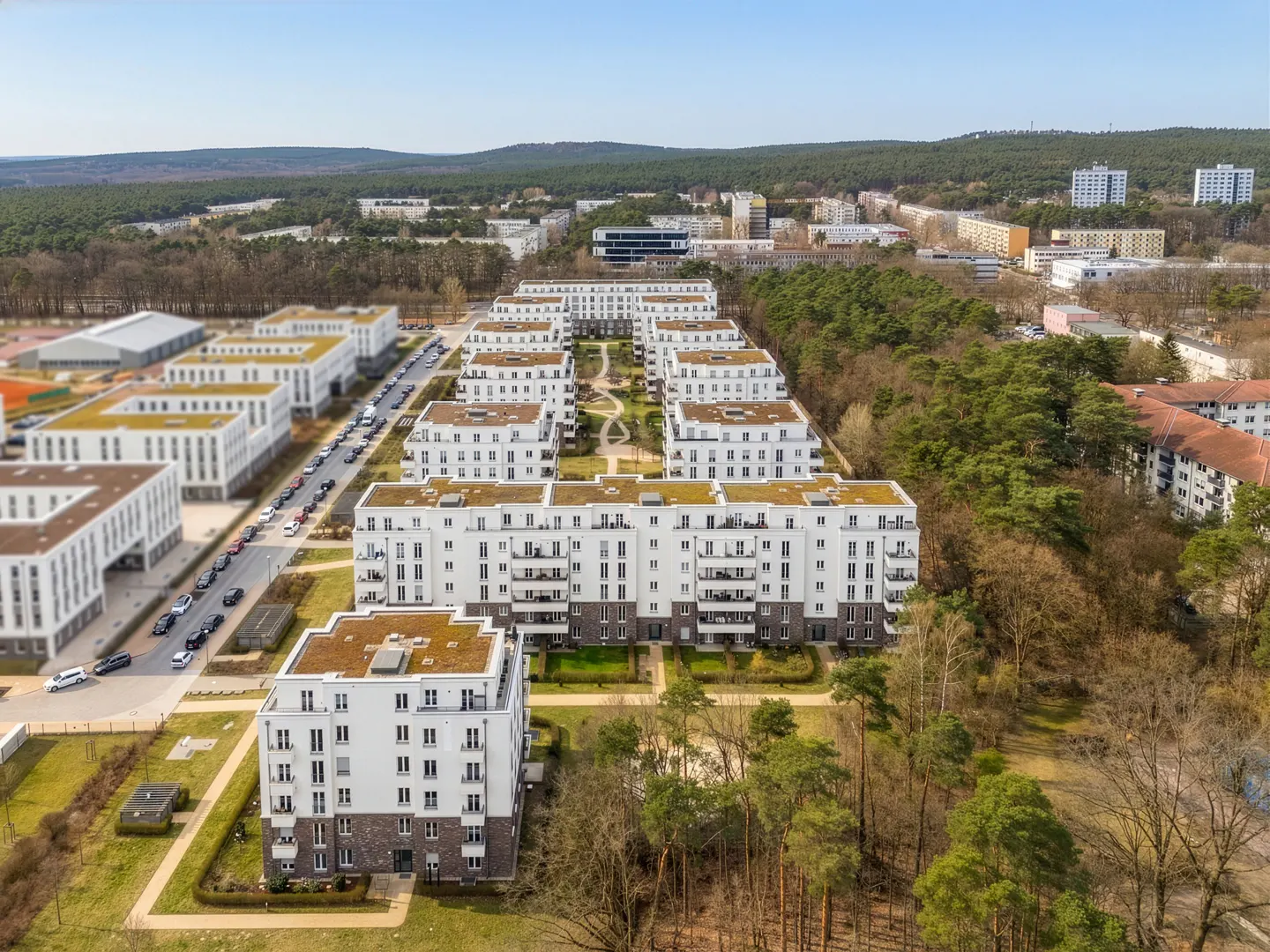 Aerial view of modern white apartment buildings with green roofs, surrounded by trees and a clear blue sky.