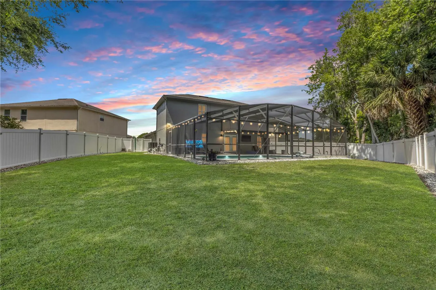 Backyard with a large green lawn, a screened-in pool with string lights, and a sunset sky with pink and blue clouds.