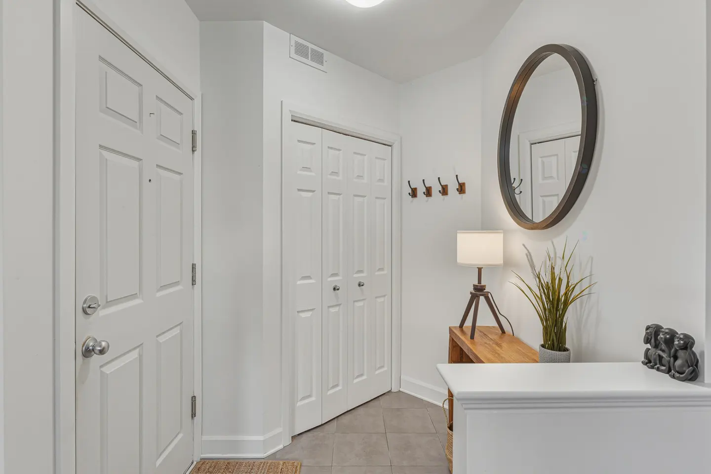 Bright entryway with white walls, gray tile floor, and white doors. A round mirror hangs above a wooden table with a lamp and plant. Hooks are on the wall.