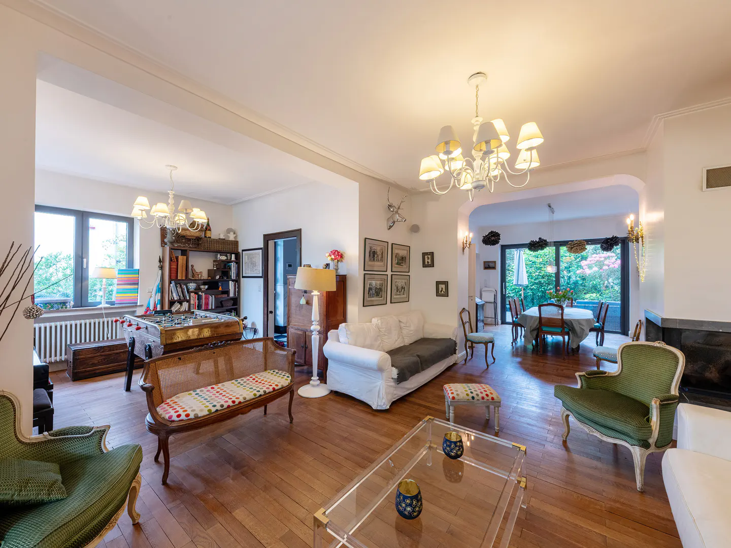 Bright living room with hardwood floors, white walls, and a chandelier. Furniture includes a sofa, chairs, and a glass coffee table. A foosball table is visible in the background.