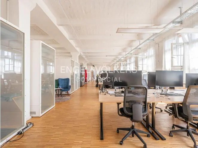 Bright, open office space with wood floors, desks, and black chairs. Glass-walled offices line the left. Windows provide natural light.