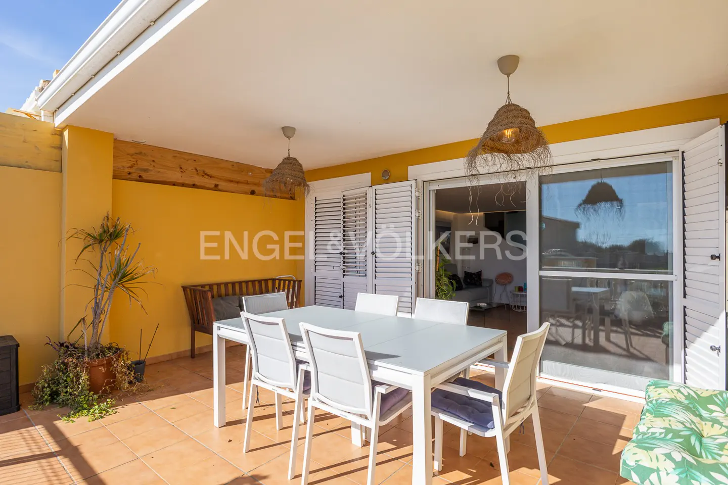 Outdoor patio with a white table, six chairs, and woven pendant lights. Yellow wall and tiled floor. Sliding glass doors lead to the interior.
